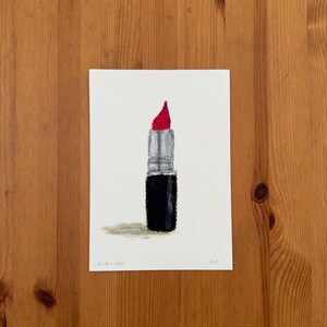 Könnte beinhalten: Eine handgezeichnete Illustration auf weißem Papier, die einen Lippenstift mit leuchtend roter Spitze, einem silbernen Metallgehäuse mit dem MAC-Logo und einem schwarzen Sockel zeigt. Der Text RUBY WOO steht unten links und BH unten rechts.