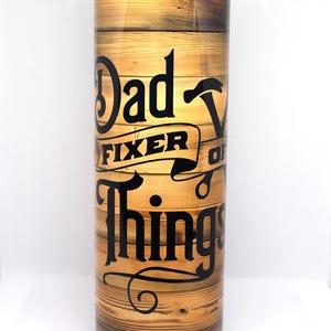 May include: A stainless steel tumbler with a wood grain design and the text "Dad - Fixer of Things" in black lettering.