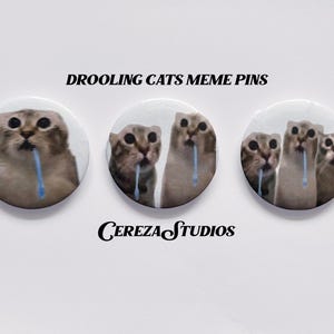 May include: Three circular pin-back buttons featuring cat memes with blue drool. The text DROOLING CATS MEME PINS and CEREZA STUDIOS is printed in black. Each button displays a different number of cats with wide eyes and blue liquid dripping from their mouths.