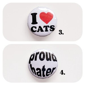 以下が含まれることがあります： 2つの丸い白いボタン。1つのボタンには赤いハートマークと「I ❤️ CATS」の文字が書かれています。もう1つのボタンには黒い文字で「proud hater」と書かれています。どちらのボタンも白い背景にあります。