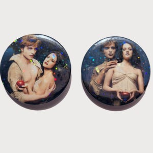 May include: Two round, black-edged buttons with a glittery overlay. Each button features a romantic image of a man and woman, one holding a red apple. The image has a dark, celestial background.