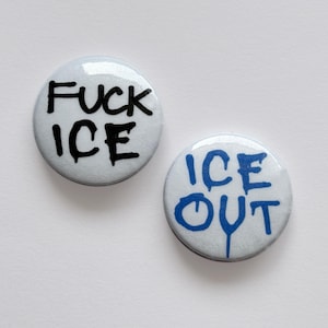 May include: Two round, white buttons with black and blue text. One button reads "FUCK ICE" in black, the other reads "ICE OUT" in blue. The buttons have a glossy finish and are set against a white background.