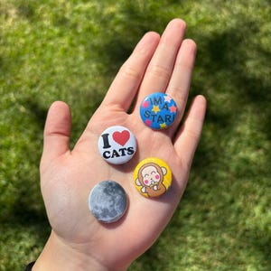 以下が含まれることがあります： 4つのカラフルなピンバックボタン。1つのボタンは灰色の月のデザインで、別のボタンは「I ❤️ CATS」と書かれており、3つ目は星付きの「I'M A STAR」と書かれており、最後は漫画の猿です。ボタンは手で持たれています。