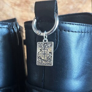 May include: A pair of black boots featuring silver-toned metal rings attached to the top pull tabs. Each ring holds a rectangular silver charm embossed with a detailed tarot card design, specifically the Wheel of Fortune, labeled La Rueda de la Fortuna.