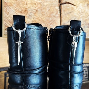 Könnte beinhalten: Ein Paar schwarze Stiefel mit silberfarbenen Metallanhängern an den oberen Zuglaschen. Der linke Stiefel zeigt einen detaillierten Schwertanhänger, während der rechte Stiefel einen Vogelschädelanhänger aufweist, die beide an silbernen Ringen hängen.