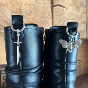 Könnte beinhalten: Zwei schwarze Lederstiefel mit silbernen Anhängern. Ein Anhänger ist ein Schwertanhänger, der andere ein Mottenanhänger. Die Anhänger hängen an den Stiefelschlaufen. Der Hintergrund ist eine Holzoberfläche.