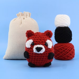 May include: A red crocheted amigurumi red panda with black eyes and white markings. The panda is sitting on a blue surface in front of a white hessian bag and three balls of yarn in white, black, and red.