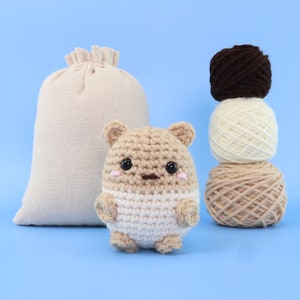 May include: A crocheted stuffed animal shaped like a bear with brown and white fur. The bear is sitting on a blue surface in front of a beige burlap bag and three balls of yarn in brown, white, and beige.