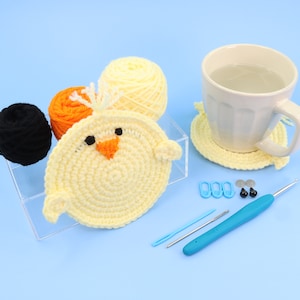 May include: A crocheted chick made with yellow, orange and black yarn. The chick is sitting on a white surface with a cup of tea, yarn and crochet hooks.
