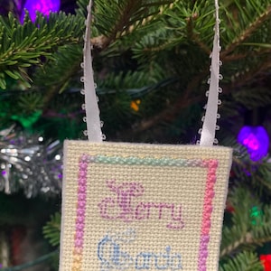 May include: A white cross-stitch ornament with a pink, yellow and green border. The ornament says "Merry Grandma and God"