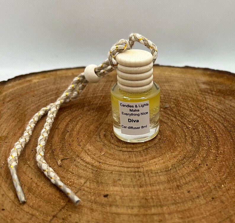 May include: A yellow glass car diffuser with a white cap and a gold string. The diffuser has the text "Candles & Lights Make Everything Nice Diva Car diffuser 8ml" printed on the label.