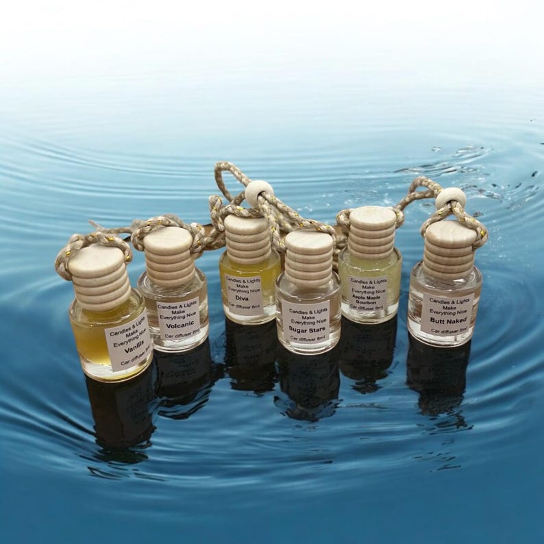 May include: Seven small glass bottles with wooden tops and string hangers, floating in blue water. Each bottle has a label with the text "Candles & Lights Make Everything Nice" and a different scent name: "Vanilla", "Volcanic", "Diva", "Sugar Stars", "Apple Maple", and "Butt Naked".