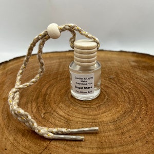May include: A small glass bottle with a wooden cap and a gold string hanging from it. The bottle is filled with a clear liquid and has a label that reads "Candles & Lights Make Everything Nice Sugar Stars Car diffuser 8ml".