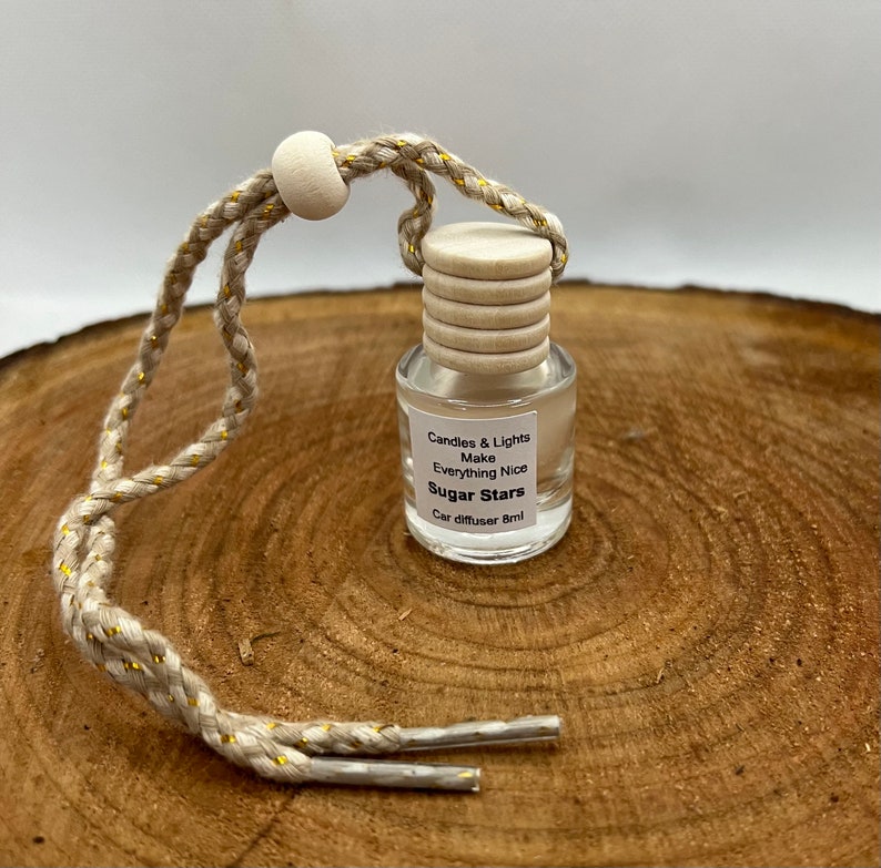 May include: A small glass bottle with a wooden cap and a gold string hanging from it. The bottle is filled with a clear liquid and has a label that reads "Candles & Lights Make Everything Nice Sugar Stars Car diffuser 8ml".