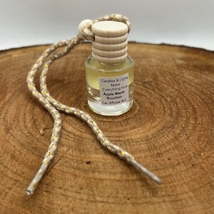 May include: A small glass bottle with a wooden cap and a gold string. The bottle contains a clear liquid and has a label that reads "Candles & Lights Make Everything Nice Apple Maple Bourbon Car diffuser 8ml".