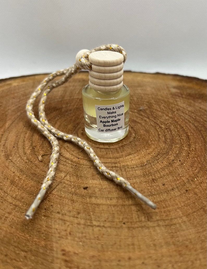 May include: A small glass bottle with a wooden cap and a gold string. The bottle contains a clear liquid and has a label that reads "Candles & Lights Make Everything Nice Apple Maple Bourbon Car diffuser 8ml".