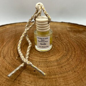 May include: A small, clear glass bottle with a wooden cap and a gold string. The bottle contains a yellow liquid and has a label that reads "Candles & Lights Make Everything Nice Vanilla Car diffuser 8ml".