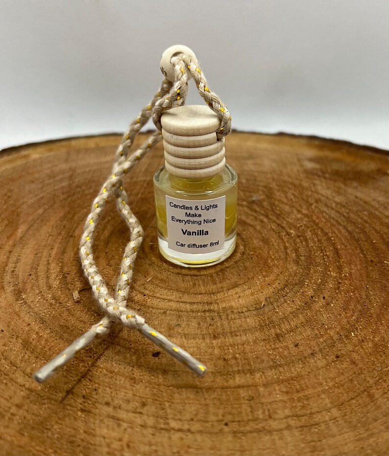 May include: A small, clear glass bottle with a wooden cap and a gold string. The bottle contains a yellow liquid and has a label that reads "Candles & Lights Make Everything Nice Vanilla Car diffuser 8ml".
