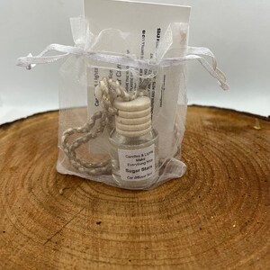 May include: A small glass bottle with a white label that reads "Candles & Lights Make Everything Nice Sugar Stars Car Diffuser Ball". The bottle is in a white organza bag with a gold cord.
