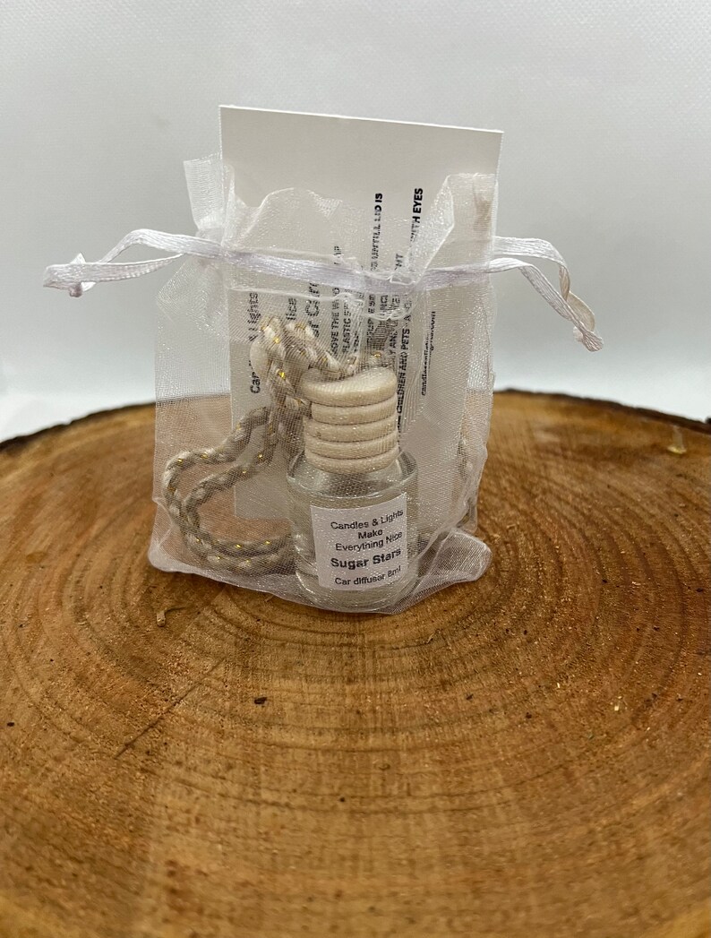 May include: A small glass bottle with a white label that reads "Candles & Lights Make Everything Nice Sugar Stars Car Diffuser Ball". The bottle is in a white organza bag with a gold cord.