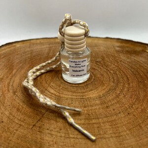 May include: A small glass bottle with a wooden cap and a beige string with gold accents. The bottle is filled with a clear liquid and has a label that reads "Candles & Lights Make Everything Nice Volcanic Car diffuser 8ml".