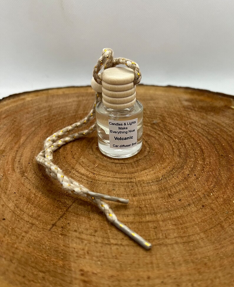 May include: A small glass bottle with a wooden cap and a beige string with gold accents. The bottle is filled with a clear liquid and has a label that reads "Candles & Lights Make Everything Nice Volcanic Car diffuser 8ml".