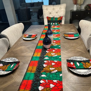 May include: A wooden dining table set with a green, red, and gold patterned table runner. The table runner has a black background and features a geometric design. There are four white plates with napkins folded on top of them. Two blue and white ceramic vases are on the table runner.