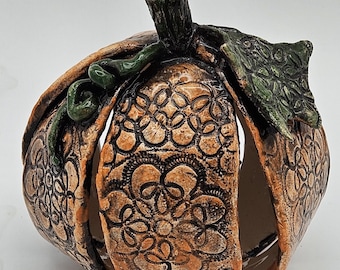 May include: A ceramic pumpkin-shaped candle holder with an open design. The pumpkin is orange with a detailed floral pattern and a green stem and leaf accents. A decorative fall-themed item.