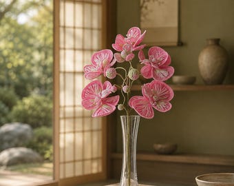 May include: A clear glass vase containing a stem of crocheted pink orchid flowers. The flowers feature intricate pink and white thread work with small green crocheted buds along the central stem, standing upright in the slender glass vessel.