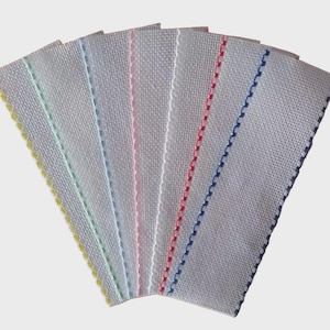 Op de afbeelding: Een set van zes rechthoekige witte stoffen stroken gerangschikt in een waaier. Elke strook is voorzien van een afzonderlijk gekleurde gestikte rand aan één zijde, waaronder gele, lichtgroene, lichtblauwe, roze, rode en donkerblauwe draden.