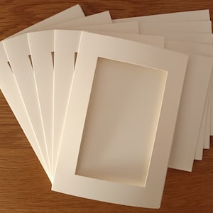 Op de afbeelding: Een stapel gebroken witte kartonnen fotomappen, elk met een rechthoekig uitgestanst venster in het midden. De mappen zijn in een waaiervormig, overlappend patroon gerangschikt, wat de gladde, matte textuur van het dikke papier benadrukt.