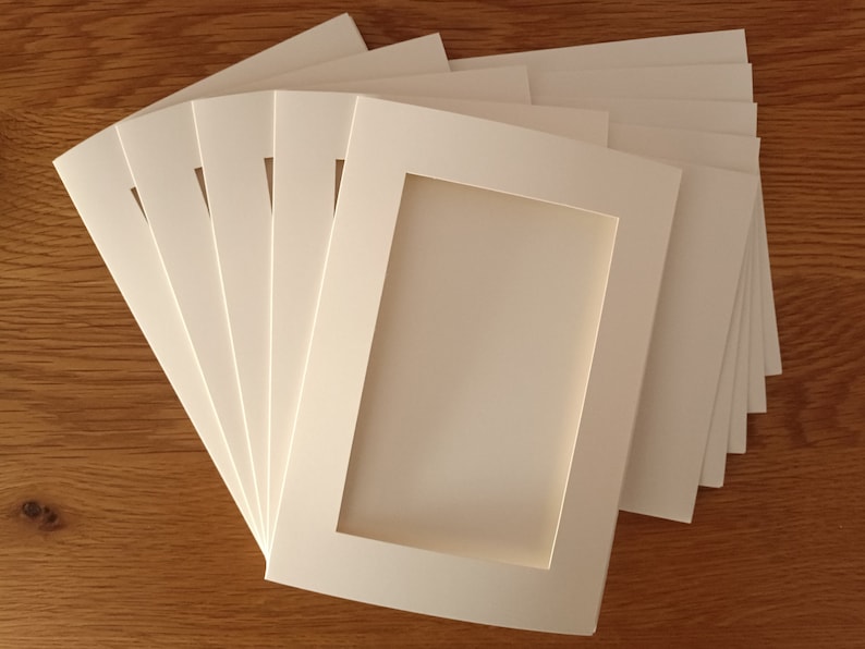 Op de afbeelding: Een stapel gebroken witte kartonnen fotomappen, elk met een rechthoekig uitgestanst venster in het midden. De mappen zijn in een waaiervormig, overlappend patroon gerangschikt, wat de gladde, matte textuur van het dikke papier benadrukt.