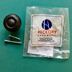 May include: Black metal cabinet knob with a textured finish and two silver screws. The knob is packaged in a clear plastic bag with a white label that reads "Hickory Hardware" and includes the company's contact information.