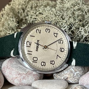 Peut inclure: Une montre-bracelet vintage avec un boîtier argenté et un cadran crème. La montre affiche des chiffres et des aiguilles noirs, avec la marque "Raketa". Elle est dotée d'un bracelet en cuir vert foncé avec des coutures blanches. La montre est posée sur de la mousse verte et des pierres.
