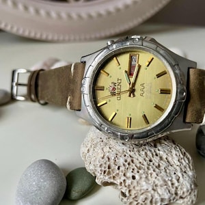 Può includere: Un orologio Orient argentato con quadrante color oro e cinturino in pelle marrone. L'orologio ha indici e lancette color oro, con un display giorno e data. L'orologio è appoggiato su una roccia strutturata di colore chiaro.