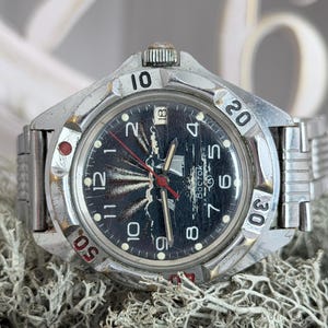 Può includere: Un orologio da polso vintage argentato con quadrante blu scuro e cinturino in acciaio inossidabile. Il quadrante presenta numeri bianchi, dettagli rossi e la parola "Восток". La lunetta ha numeri da 10 a 50. L'orologio è su uno sfondo strutturato.