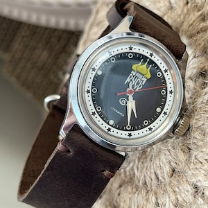 Puede incluir: Un reloj de pulsera plateado con una esfera negra y una correa de cuero marrón. La esfera del reloj presenta un círculo blanco con marcas negras y una estrella blanca en cada marca de hora. El centro de la esfera presenta una imagen amarilla y roja de una iglesia con la palabra "РФСФ" en letras rojas.