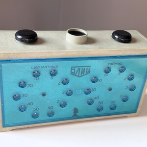 May include: Vintage chess timer with a light blue face and a wooden frame. The timer has two black buttons on top and a series of buttons and numbers on the face. The text on the face is in a foreign language.