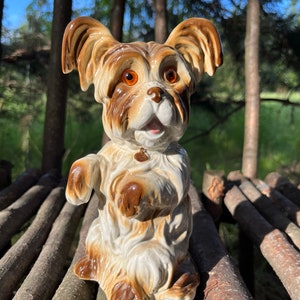 Könnte beinhalten: Eine Keramik-Hundefigur in Braun- und Weißtönen. Der Hund ist mit erhobener Pfote und einem kleinen Halsbandanhänger dargestellt. Die Figur hat große Augen und steht vor einem natürlichen Hintergrund aus Bäumen und Baumstämmen.