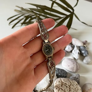 May include: Vintage gold-toned watch with an oval black watch face and ornate filigree detailing. The watch features a decorative band with intricate patterns and small gemstones. The watch is held in a hand, with a background of rocks and greenery.