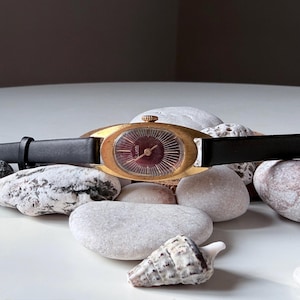 Puede incluir: Reloj de pulsera vintage dorado con esfera granate y correa de cuero negro. El reloj tiene una esfera ovalada y se encuentra sobre un fondo de piedras lisas y claras y una concha. Un reloj clásico.