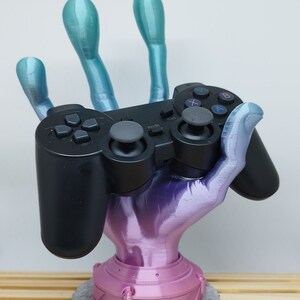 Könnte beinhalten: Eine 3D-gedruckte Hand hält einen schwarzen Videospiel-Controller. Die Hand ist in einem Farbverlauf von Blau und Rosa bemalt. Der Controller hat Knöpfe mit den Beschriftungen 'Start', 'Mode', 'A', 'B', 'X' und 'Y'. Die Hand steht auf einem rosa Sockel mit einem grauen Filzpad darunter.