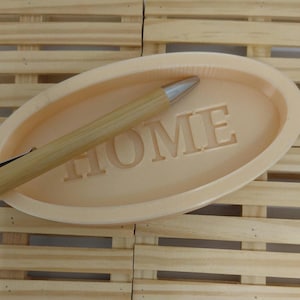 May include: A light peach-colored oval dish with the word "HOME" embossed in the center. A wooden pen with a silver tip rests on the dish. The dish and pen are set against a light wooden background.