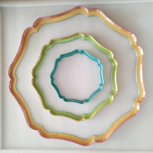 Op de afbeelding: Drie decoratieve schalen met gegolfde randen. De grootste schaal is goud met een roze rand, de middelste schaal is groen en de kleinste schaal is teal.