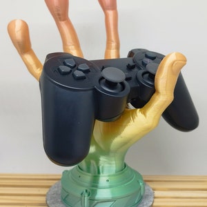 Könnte beinhalten: Eine 3D-gedruckte Hand hält einen schwarzen Videospiel-Controller. Die Hand ist in einem Farbverlauf von Pink, Orange und Gelb bemalt. Die Basis der Hand ist grün und steht auf einem grauen runden Sockel.