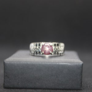 Peut inclure: Une bague en argent avec une pierre précieuse rose en forme d'étoile sertie au centre. La bande est décorée d'un motif géométrique.