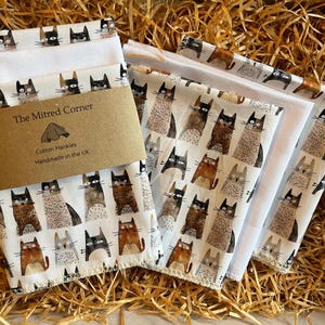 May include: A collection of white cotton hankies with a repeating pattern of various cat illustrations in brown, black, and gray. The hankies are packaged with a brown paper label that reads "The Mitred Corner" and "Cotton Hankies Handmade in the UK."