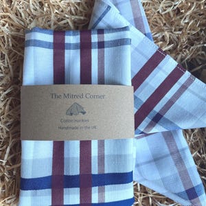May include: A folded cotton handkerchief featuring a white, burgundy, and navy blue plaid pattern. A brown paper band wraps around the centre, printed with the text The Mitred Corner, a graphic icon of a folded cloth, and Cotton Hankies Handmade in the UK.