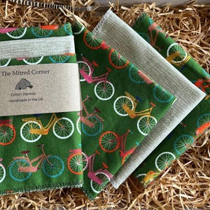 May include: A collection of cotton hankies featuring a bicycle design in green, orange, pink, and yellow. The hankies are packaged with a brown paper label that reads "The Mitred Corner" and "Cotton Hankies Handmade in the UK."