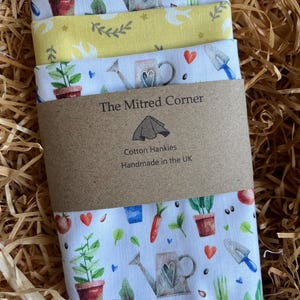 Può includere: Una pila di quattro fazzoletti di cotone con motivi a tema giardino. Il fazzoletto superiore è giallo con foglie verdi, mentre gli altri presentano annaffiatoi, piante e verdure. I fazzoletti sono avvolti con una fascia di carta marrone con la scritta "The Mitred Corner" e "Cotton Hankies, Handmade in the UK."